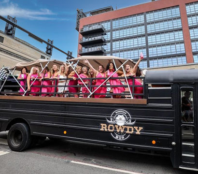 Group of women—mostly in bright pink dresses with one in white—cheering on an open-top party bus rolling through a downtown city street under a blue sky.