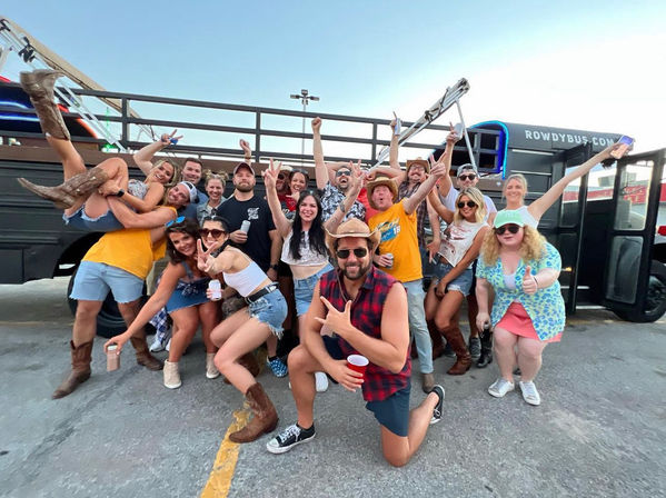 Energetic group of friends cheering and posing in front of a party bus in a parking lot, wearing summer outfits and cowboy boots, holding drinks and phones.