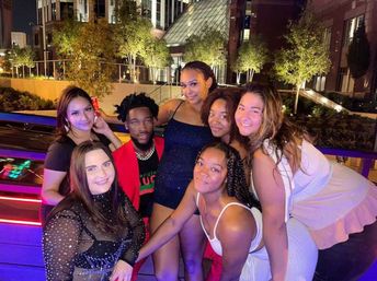 Seven friends posing on an outdoor rooftop terrace at night, colorful neon and purple lights, dressed in party outfits and smiling together with illuminated city buildings and trees in the background.