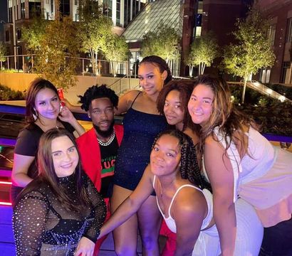 Seven friends posing on an outdoor rooftop terrace at night, colorful neon and purple lights, dressed in party outfits and smiling together with illuminated city buildings and trees in the background.