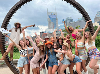 Cheerful group of young women in summer outfits and cowboy hats posing inside a large rusted circular gear sculpture with the Nashville, Tennessee skyline and twin‑spired tower in the background.