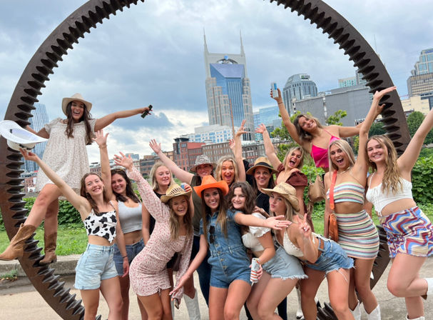 Cheerful group of young women in summer outfits and cowboy hats posing inside a large rusted circular gear sculpture with the Nashville, Tennessee skyline and twin‑spired tower in the background.