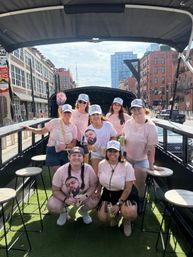 Bachelorette party crew in matching pink 'let's go girls' shirts and white trucker hats posing with canned drinks and cardboard face cutouts on an open-air party wagon down a downtown street of brick buildings and a glass high-rise.