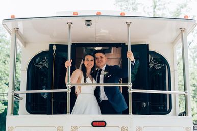 Smiling bride in a white gown and groom in a navy suit embrace on the rear platform of a white wedding trolley, holding chrome railings with leafy green trees in the background.
