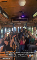 Group of friends smiling and waving inside a wood-paneled Manhattan trolley on an NYC pizza tour, sunlit windows and festive ceiling lights