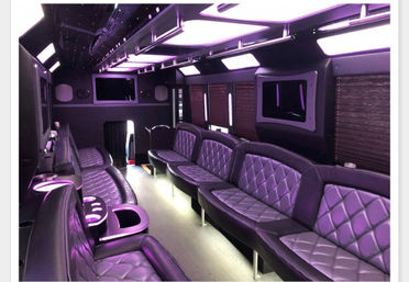 Interior of a luxury party bus with purple LED ambient lighting, quilted leather bench seats lining both sides, cup holders, overhead light panels and window shades — spacious mobile lounge for nightlife and events.