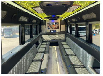Luxury party bus interior with parallel quilted silver-and-black bench seating, wood-look aisle, patterned green LED ceiling, overhead speakers and rear flat-screen TV, parked shuttle visible through side windows.