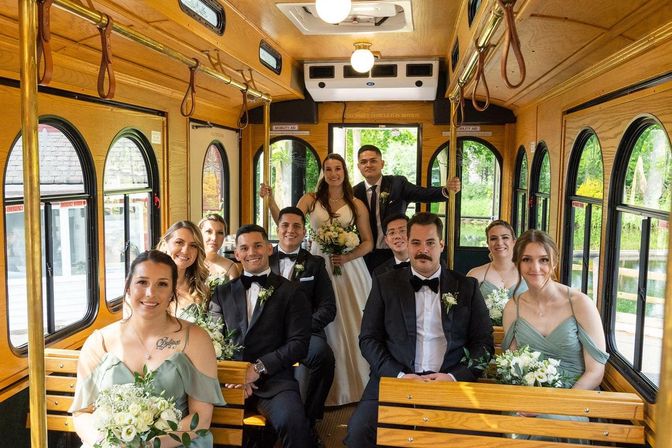 Wedding party on a vintage wooden trolley — bride in a white gown at center, bridesmaids in sage-green dresses and groomsmen in black tuxes holding bouquets, sitting on wooden benches with arched windows showing green trees outside.