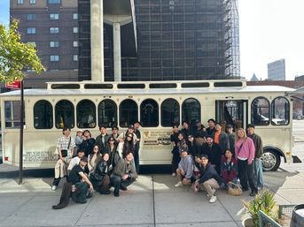 Large group of people smiling and posing in front of a cream vintage-style tour trolley parked on a sunny downtown sidewalk, ready for a city pizza tour.