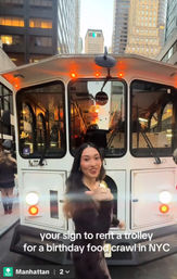 Woman gesturing in front of a white vintage-style trolley on a Manhattan street at dusk, skyscrapers and warm interior lights behind — playful scene suggesting a NYC birthday food crawl.