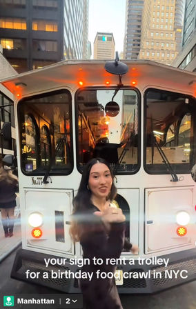 Woman gesturing in front of a white vintage-style trolley on a Manhattan street at dusk, skyscrapers and warm interior lights behind — playful scene suggesting a NYC birthday food crawl.