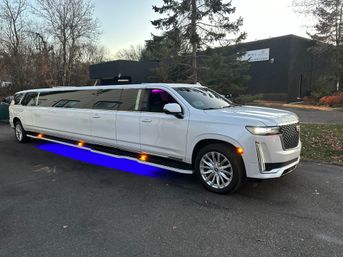 Sleek white stretch SUV limousine with blue LED underglow parked in a suburban lot at dusk, trees and a low-rise commercial building visible in the background.