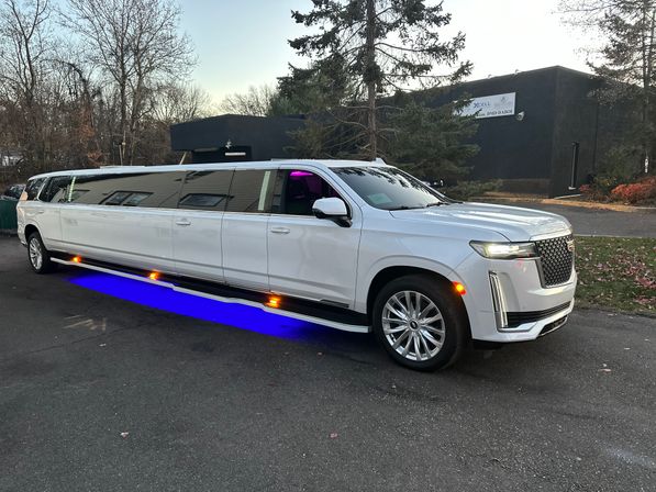 Sleek white stretch SUV limousine with blue LED underglow parked in a suburban lot at dusk, trees and a low-rise commercial building visible in the background.