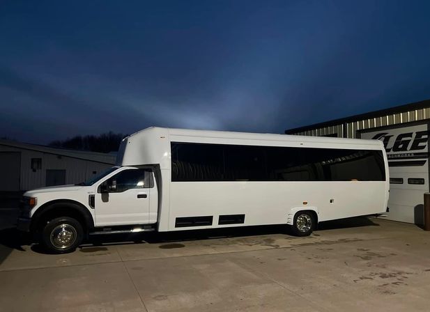 Sleek white stretch party bus parked outside a metal garage on a concrete lot at dusk, long tinted windows and chrome wheels