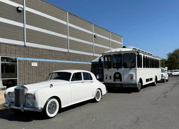 Classic white Rolls-Royce sedan and matching white trolley bus parked outside a beige industrial building in a sunny commercial parking lot