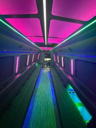 Interior of a luxury party bus with neon pink ceiling panels, green and blue LED strip lights, long quilted bench seating and a glowing central aisle