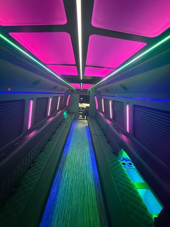 Interior of a luxury party bus with neon pink ceiling panels, green and blue LED strip lights, long quilted bench seating and a glowing central aisle