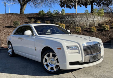 White Rolls‑Royce luxury sedan parked on sunny suburban pavement, chrome grille and Spirit of Ecstasy hood ornament, stone retaining wall and trees in the background.