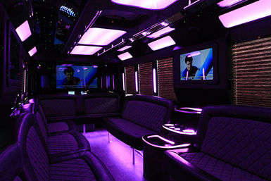 Lavish purple-lit party bus interior with quilted leather bench seating, overhead LED panels, wall-mounted TVs and illuminated cup holders — VIP nightlife ride.