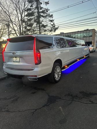 White stretched Cadillac Escalade SUV limousine with blue underglow and amber side lights parked on an urban street at dusk, bare trees and office building reflected in tinted windows.