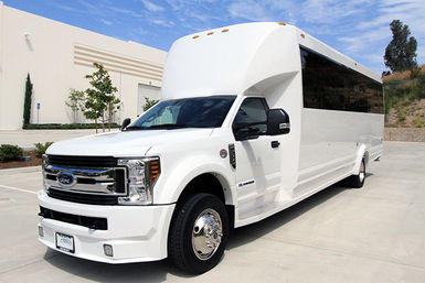 White Ford-based luxury shuttle bus parked in a sunny commercial parking lot beside a modern warehouse under a bright blue sky