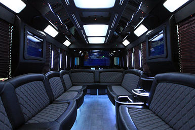 Luxury party bus interior with quilted leather wraparound bench seating, ambient blue LED floor and ceiling lighting, wall-mounted TVs and dark wood accents