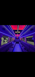 Vibrant party bus interior with purple and pink LED ceiling panels, quilted side bench seating, skylight window, and striped aisle floor.