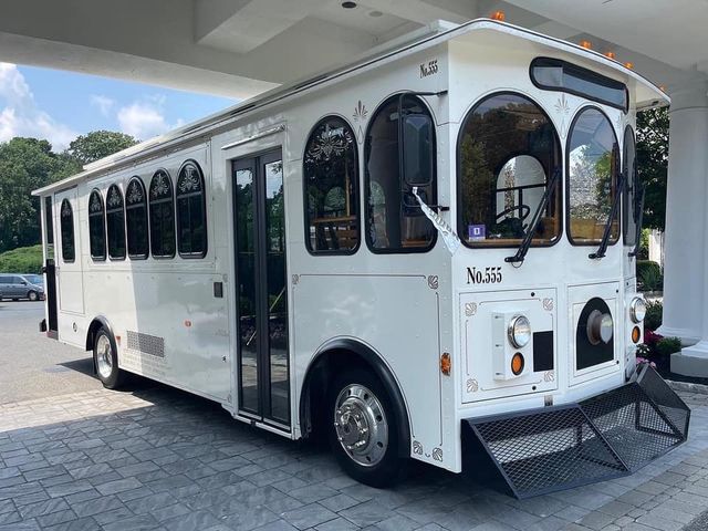 White vintage-style shuttle trolley parked under a hotel porte-cochere — arched passenger windows, glass double doors, chrome wheels and front fold-down ramp.