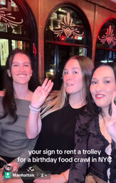 Three smiling friends waving inside a wooden Manhattan trolley at night on a birthday food crawl in NYC, with arched decorative windows and warm red lighting.