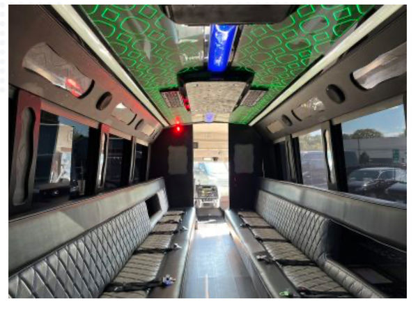 Luxury party bus interior with long quilted black leather bench seats lining both sides, green patterned LED ceiling lights with blue accents, overhead panels and seatbelts, windows showing a sunny parking lot outside.