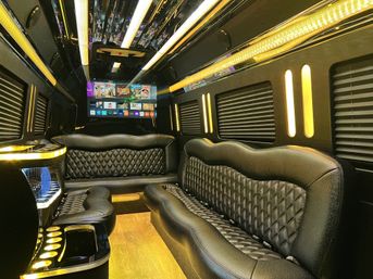 Luxurious party bus interior with quilted black leather bench seats, warm yellow LED accent lighting, wood-look floor, cup holders and wall-mounted flat-screen TV.