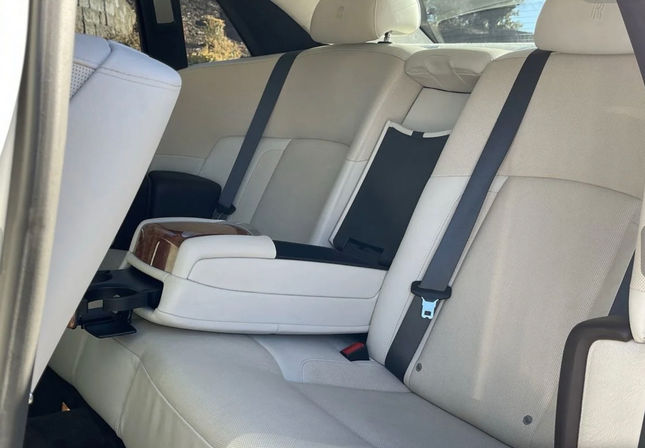 Beige leather rear car seats with folded center armrest, wood-trim cupholder, black seat belts and headrests — clean, plush sedan interior.