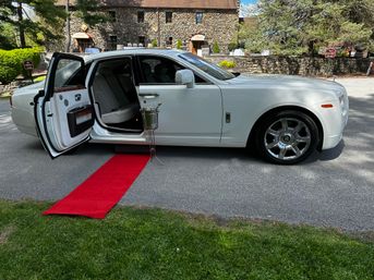 White luxury sedan with open rear door, red carpet runner and champagne bucket on a stand outside a stone event venue on a tree-lined driveway.