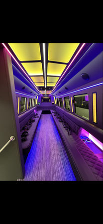 Luxury party bus interior with long quilted bench seating, central aisle, carpeted floor and vibrant purple and yellow LED ceiling and accent lighting with built-in cup holders.