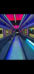 Spacious luxury party-bus interior with long quilted bench seats on both sides, neon pink and blue LED ceiling panels, illuminated floor aisle, seatbelts and cup holders — modern limo-style shuttle vibe.