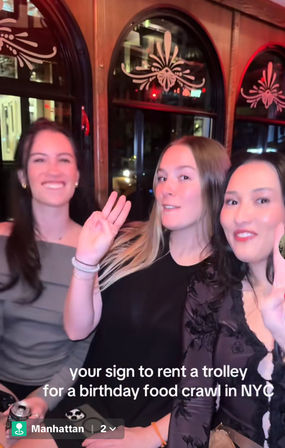 Three friends smiling and waving inside a vintage Manhattan trolley with decorative arched windows — caption reads 'your sign to rent a trolley for a birthday food crawl in NYC'.