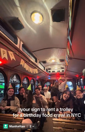 Smiling group wearing party hats inside a decorated Manhattan trolley at night, wooden bench seats, bunting and warm lights for a birthday food crawl — overlay text about renting a trolley in NYC.
