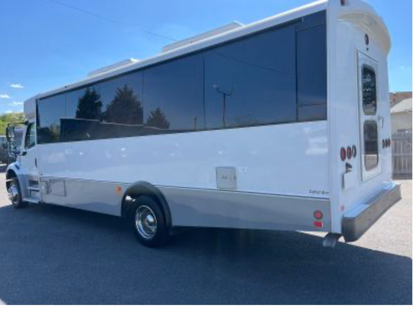 Sleek white passenger shuttle bus with large dark tinted windows and rear entry door parked on a sunny asphalt lot under a blue sky