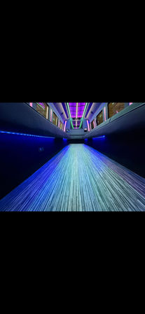 Low-angle view inside a stretch limousine showing a long striped illuminated aisle, black leather bench seating, and vibrant purple, teal and blue LED ceiling and under-seat lighting for a luxe party vibe.