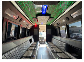 Spacious party bus interior with long black quilted leather bench seats facing a central aisle, green patterned LED ceiling lights, blue and red accent lighting, large windows and visible seatbelts.