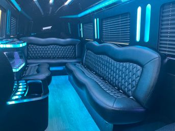 Sleek luxury party bus interior with black quilted leather bench seats, blue LED accent lighting and starry ceiling lights, wood-look floor, built-in cup holders and privacy blinds.