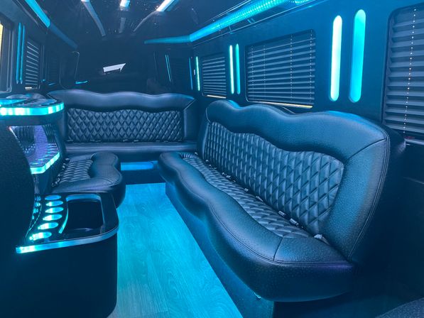 Sleek luxury party bus interior with black quilted leather bench seats, blue LED accent lighting and starry ceiling lights, wood-look floor, built-in cup holders and privacy blinds.