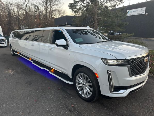 White stretched Cadillac Escalade SUV limousine with blue underglow and amber side lights, parked on asphalt near trees — luxury party/airport limo.