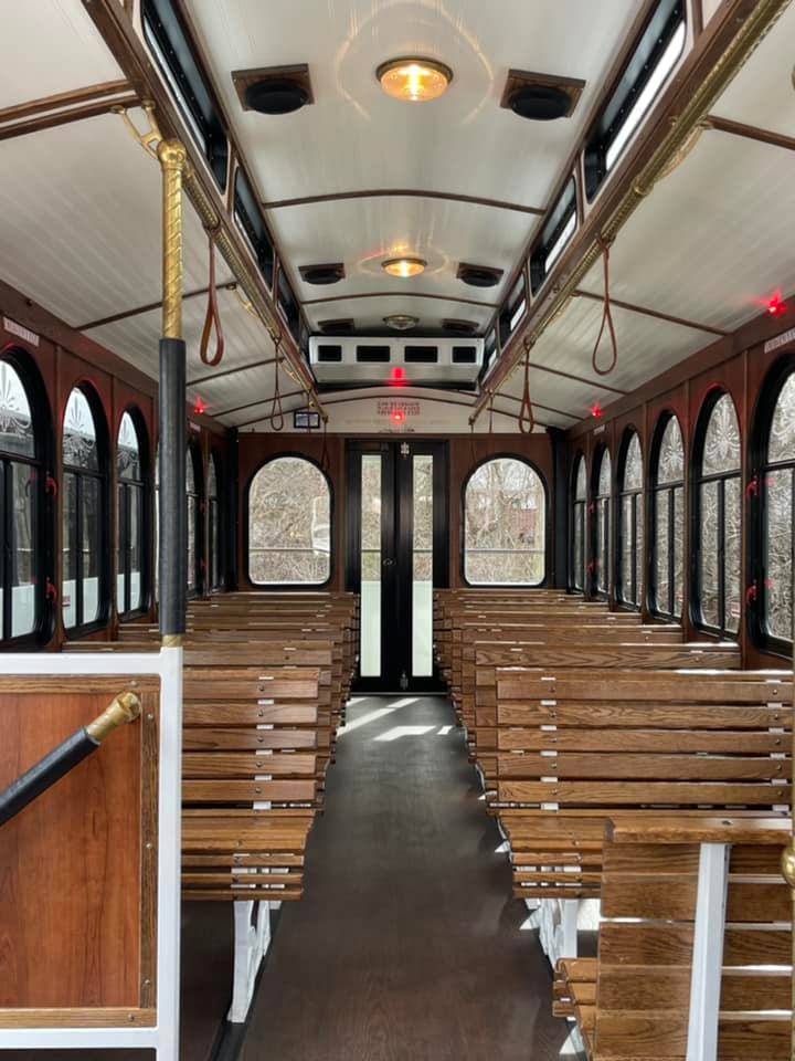 Sunlit vintage trolley interior with rows of wooden bench seats, arched windows, brass poles and leather hand straps flanking a central aisle