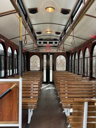 Sunlit vintage trolley interior with rows of wooden bench seats, arched windows, brass poles and leather hand straps flanking a central aisle