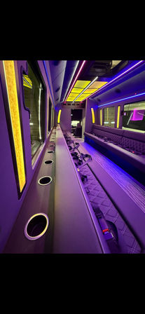 Party bus interior with purple and yellow LED ambient lighting, long quilted bench seating, multiple cup holders and visible seat belts.