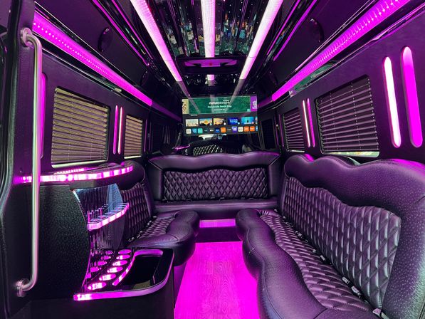 Glam party limousine interior with quilted black leather bench seats, curved cocktail bar and cup holders, mirrored ceiling, and vibrant pink-purple LED strip lighting framing a large flat-screen TV.