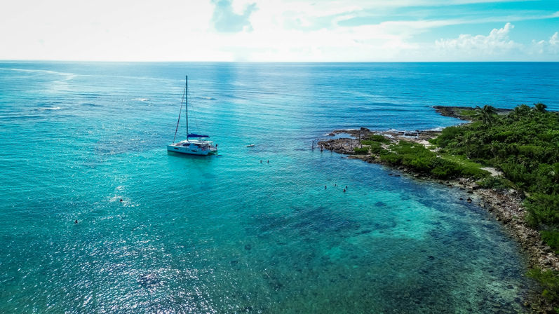 White catamaran anchored in turquoise tropical waters off a palm‑fringed rocky coastline, with snorkelers in the clear blue sea and a bright sky on the horizon.