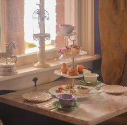 Cozy window-side afternoon tea in a café: three-tiered stand of pastries, finger sandwiches and desserts on a marble table with a floral teacup, decorative birdcage and carousel figurine in warm soft light.