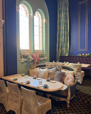 Cozy Victorian-style tea room with arched leaded windows, deep blue walls, ornate gold-trim sofa, pastel teacups on a set table and floral window decor.
